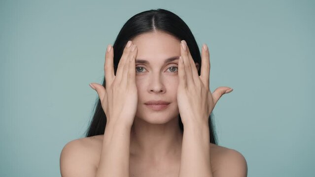 Young brunette woman without clothes touches the smooth, healthy skin of her face. Close up portrait of a young woman isolated on a blue background. Natural beauty, cosmetology, skin care. Slow motion