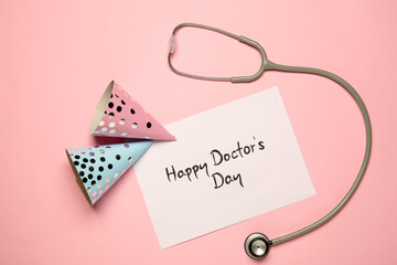 Card with phrase Happy Doctor's Day, stethoscope and party hats on pink background, flat lay