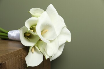 Beautiful calla lily flowers tied with ribbon on wooden table near olive wall, closeup. Space for text