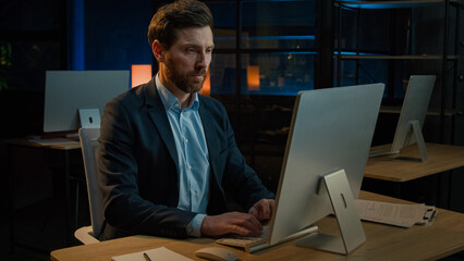 Male business portrait confident focused businessman millennial caucasian senior 40s adult man chief boss leader sit at desk with computer search online data working alone in evening office indoors