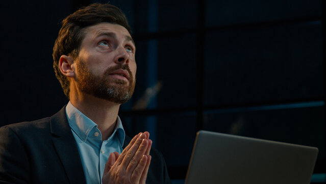 Hopeful dreaming Caucasian middle aged businessman adult man sit with computer at evening dark office ask god bless help praying hope say prayers please wish luck to get good result believe fortune