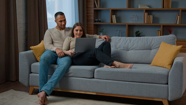 Diverse multiracial couple sitting on sofa surfing online website on laptop using computer at home relaxing woman and man talking buying retail goods shopping online make order spend weekend on couch