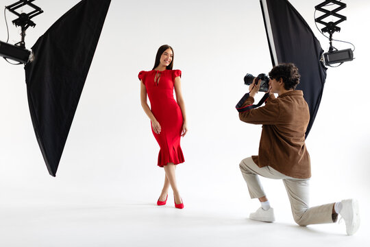 Professional young man photographer capturing portraits of elegant lady in red dress, woman posing on white background