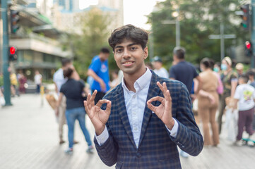 A stylish guy in a blue checkered suit is holding up a two 'okay' sign and smiling while standing outside, in the city. Building, people, stop lights and trees are shown in the background.