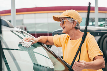 Smiling senior woman in yellow t-shirt and hat cleans the car in a self-service car wash station...