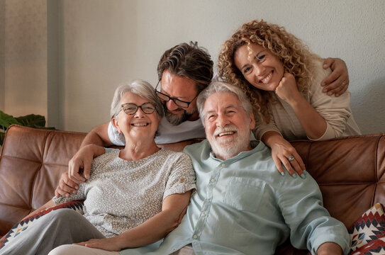 Happy Multigenerational Family Group Sitting On Sofa At Home While Spending Time Together. Four Handsome Bonding People, Two Generations Looking At The Camera