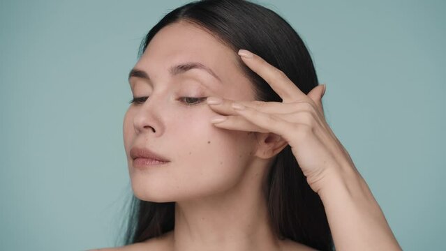 A seminude woman massages the skin around the eyes. A woman struggles with mimic wrinkles by gently massaging the skin under her eyes. Portrait of a woman on a blue background close up. Slow motion.