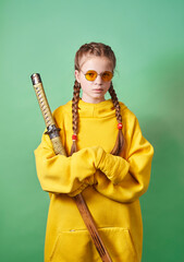 A girl in a yellow hoodie with a large Japanese sword in a wooden scabbard on a green background....