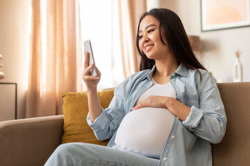 Happy Pregnant Korean Lady Using Smartphone Sitting At Home