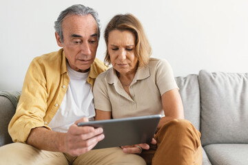 Puzzled senior spouses using digital tablet having hard time learning or browsing internet, sitting...