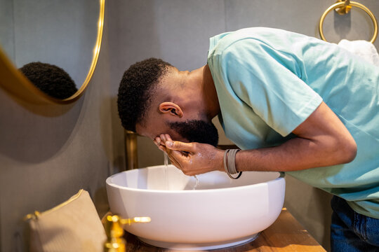 Tired sleepy African American guy washing face with water in morning, black man standing in bathroom doing daily hygiene routine at home before going to work. Facial cleansing for dark skin