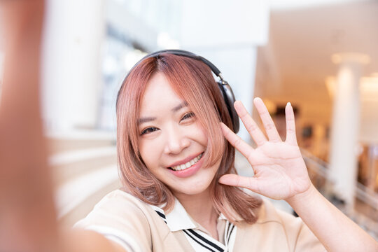 happy female asian student selfie and video conference with her high school friend by technology smart phone and wireless headphone for listen music after test exam in library campus in university