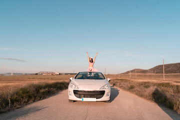 white European girl sits in the seat of her white convertible car and raises her hands to the sky...