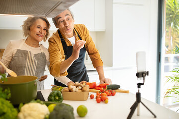 Happy aged caucasian couple famous bloggers cooking healthy dinner and streaming on cellphone in kitchen interior