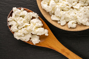 Fresh cottage cheese in ceramic plate and wooden spoon on slate stone, macro, top view.