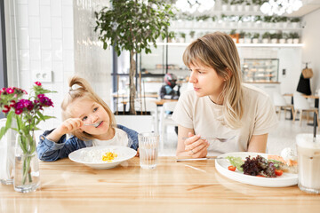 An attractive mother and her pretty girl, together have breakfast rest in the cafe at the table,...