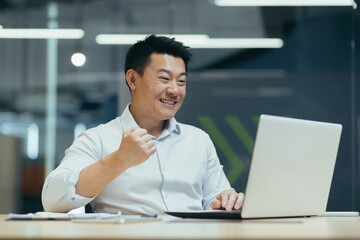 Young Asian businessman working in office at laptop and happy with success, showing victory hand gesture, satisfied with result, successful deal.