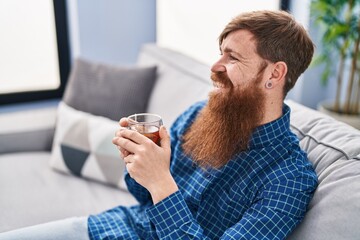 Young redhead man drinking tea sitting on sofa at home