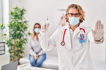 Naklejka premium Blond man wearing doctor uniform and medical mask holding syringe with open hand doing stop sign with serious and confident expression, defense gesture