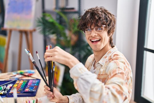 Young hispanic man holding paintbrushes sitting on desk at art studio