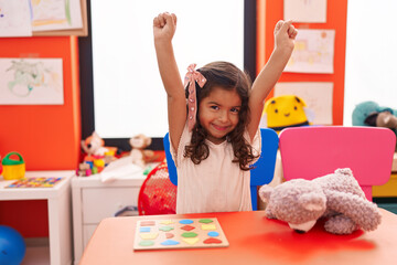 Adorable hispanic girl playing with maths puzzle game doing winner gesture at kindergarten