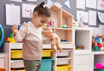 Adorable hispanic girl playing with baby doll standing at kindergarten