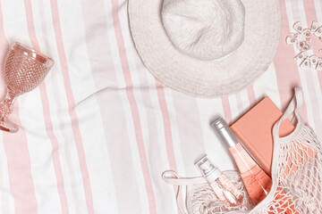 Summer flat lay with sun hat, cosmetics product, bottle rose wine, book in mesh bag on stripes beach towel as background. Summer beach holiday concept. Summertime relax, lifestyle photo