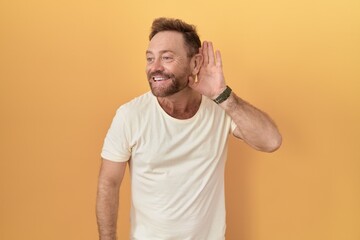 Middle age man with beard standing over yellow background smiling with hand over ear listening an hearing to rumor or gossip. deafness concept.