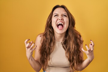 Young hispanic woman standing over yellow background crazy and mad shouting and yelling with aggressive expression and arms raised. frustration concept.