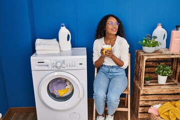 Young latin woman drinking coffee waiting for washing machine at laundru room