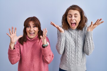 Mother and daughter standing over blue background celebrating mad and crazy for success with arms...