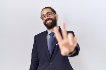 Hispanic man with beard wearing suit and tie smiling looking to the camera showing fingers doing victory sign. number two.