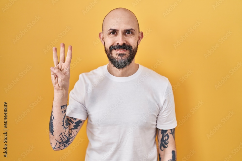 Wall mural Young hispanic man with tattoos standing over yellow background showing and pointing up with fingers number three while smiling confident and happy.