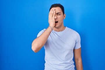 Fototapeta premium Young hispanic man standing over blue background yawning tired covering half face, eye and mouth with hand. face hurts in pain.