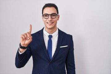 Young hispanic man wearing suit and tie showing and pointing up with finger number one while smiling confident and happy.
