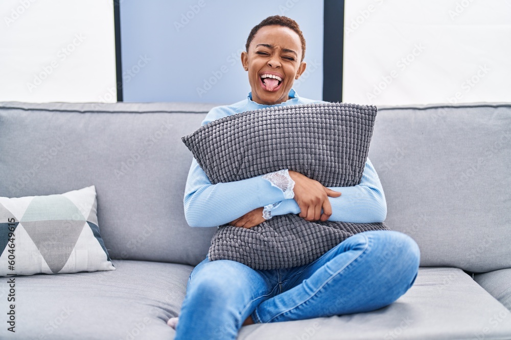 Sticker african american woman sitting on the sofa at home hugging pillow sticking tongue out happy with fun