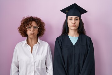Hispanic mother and daughter wearing graduation cap and ceremony robe puffing cheeks with funny...