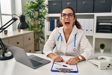 Young hispanic woman wearing doctor uniform and stethoscope angry and mad screaming frustrated and furious, shouting with anger. rage and aggressive concept.