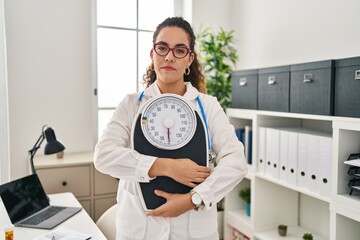 Young hispanic woman working at dietitian clinic relaxed with serious expression on face. simple...