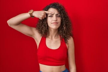 Hispanic woman with curly hair standing over red background pointing unhappy to pimple on forehead,...