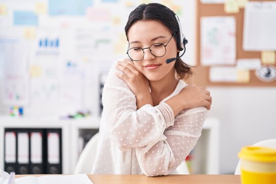 Hispanic young woman working at the office wearing headset and glasses hugging oneself happy and positive, smiling confident. self love and self care