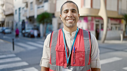 Young hispanic man volunteer smiling wearing vest at street - obrazy, fototapety, plakaty