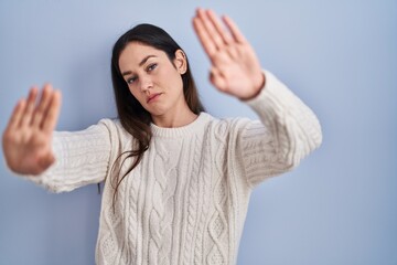 Young brunette woman standing over blue background doing frame using hands palms and fingers, camera perspective