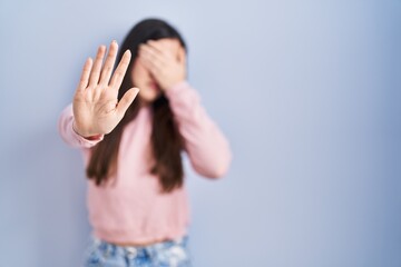 Obraz na płótnie Canvas Young brunette woman standing over blue background covering eyes with hands and doing stop gesture with sad and fear expression. embarrassed and negative concept.