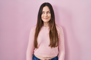 Young brunette woman standing over pink background smiling looking to the side and staring away thinking.