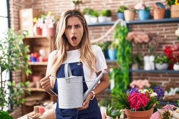 Young blonde woman working at florist shop in shock face, looking skeptical and sarcastic, surprised with open mouth