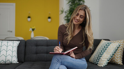 Young blonde woman writing on notebook sitting on sofa at home