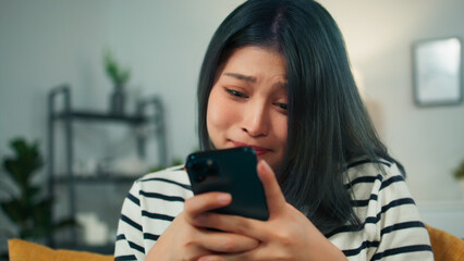 Beautiful Asian girl happy holding smartphone checking SMS e-mail received good news feeling surprised and excited. Happy young woman smile looking at smartphone sitting on couch in modern living room