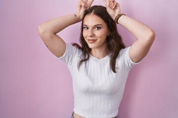 Young hispanic girl standing over pink background posing funny and crazy with fingers on head as bunny ears, smiling cheerful