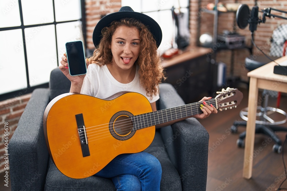 Poster young caucasian woman playing classic guitar at music studio holding smartphone sticking tongue out 